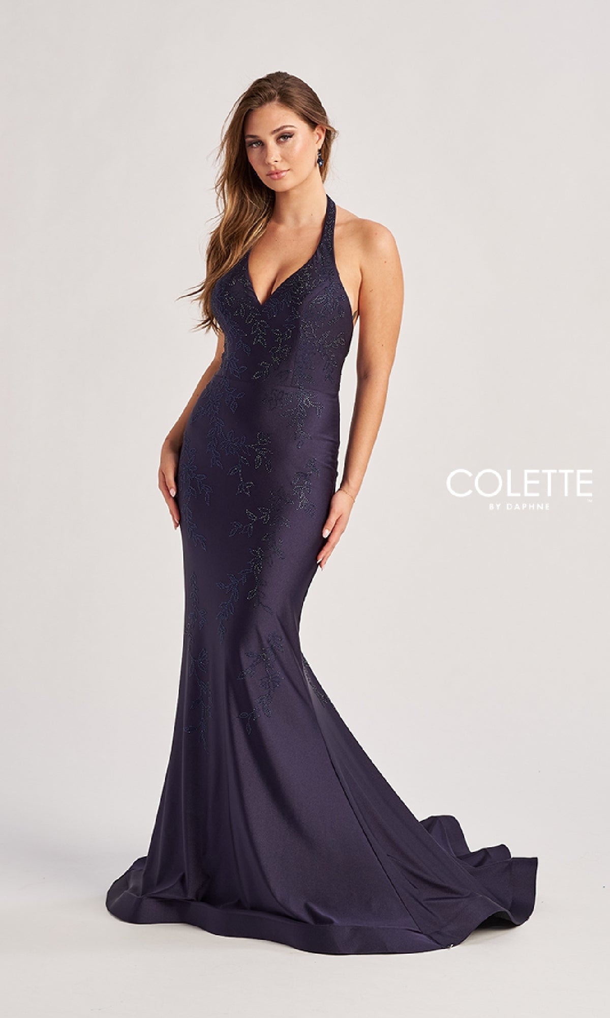 A woman poses against a plain light background wearing the Colette CL8580 long prom dress—a fitted, floor-length dark blue mermaid gown with a halter v-neckline and subtle embellishments.